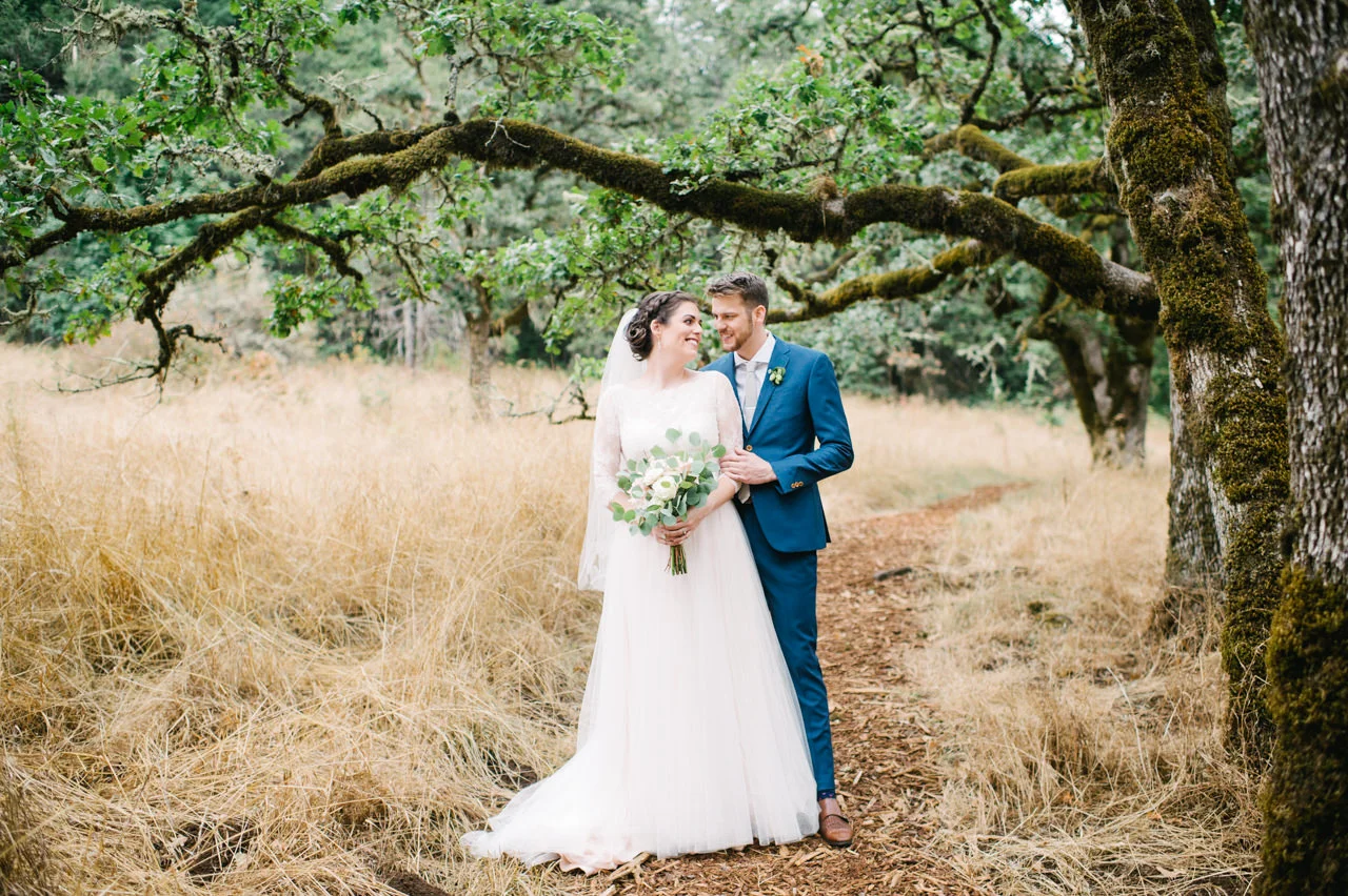 A bride and groom smiling at each other beneath a moss-covered oak in tall golden grass at Mount Pisgah, Oregon