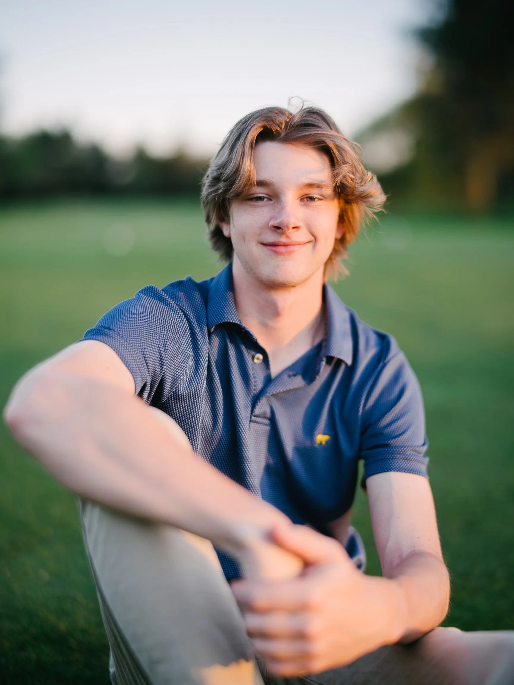 A high school senior sitting on a lawn at sunset, arms resting on one raised knee, wearing a blue polo shirt
